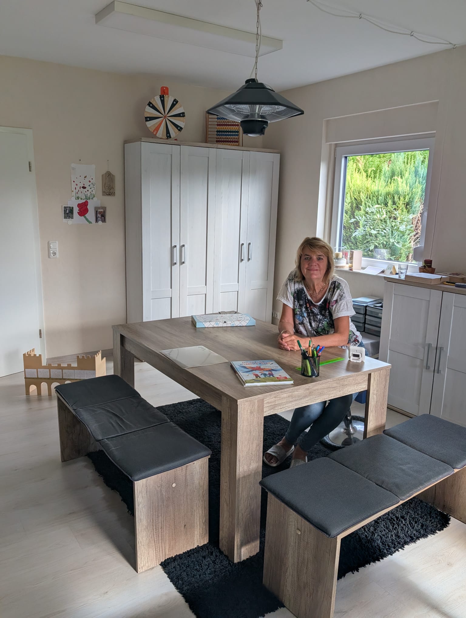 Der Therapieraum: Judith Behnke sitzt an einem großen Holztisch mit Sitzbänken, dahinter ein heller Schrank, ein Abakus und Lernspiele.
