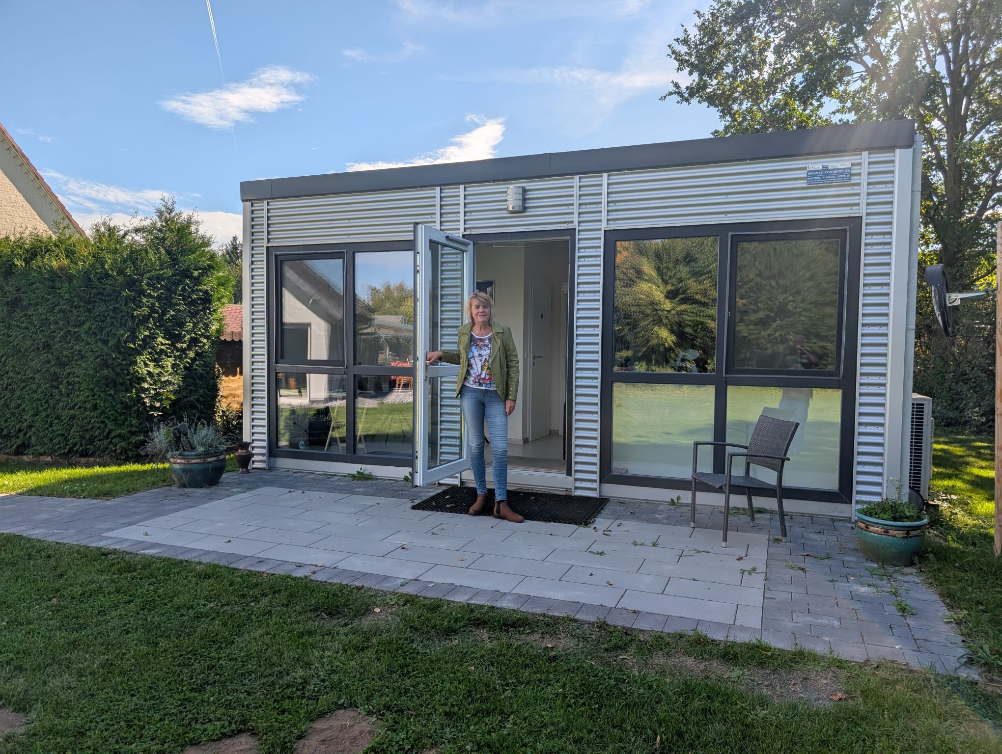 Judith Behnke steht in der geöffneten Eingangstür der Praxis, davor eine gepflasterte Terrasse mit Stuhl und Pflanzkübeln.