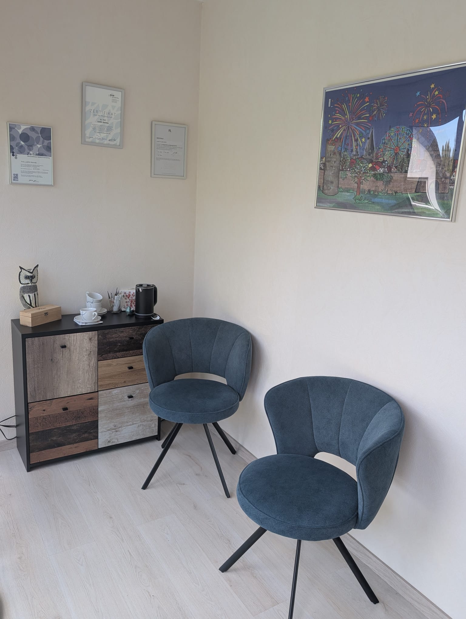 Eine Sitzecke mit zwei blauen Sesseln, einem Sideboard mit Teetassen und Wasserkocher sowie gerahmten Zertifikaten an der Wand.